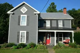 Gray two-story house with white trim, red door, and covered porch, surrounded by greenery under a blue sky