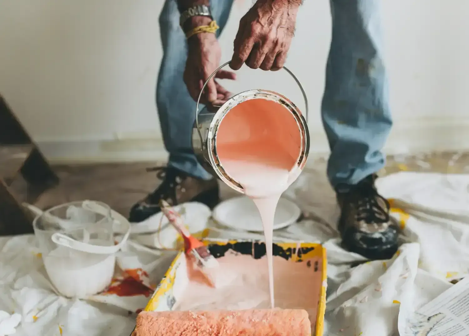 Person pours white paint into an orange paint tray on a drop cloth-covered floor.