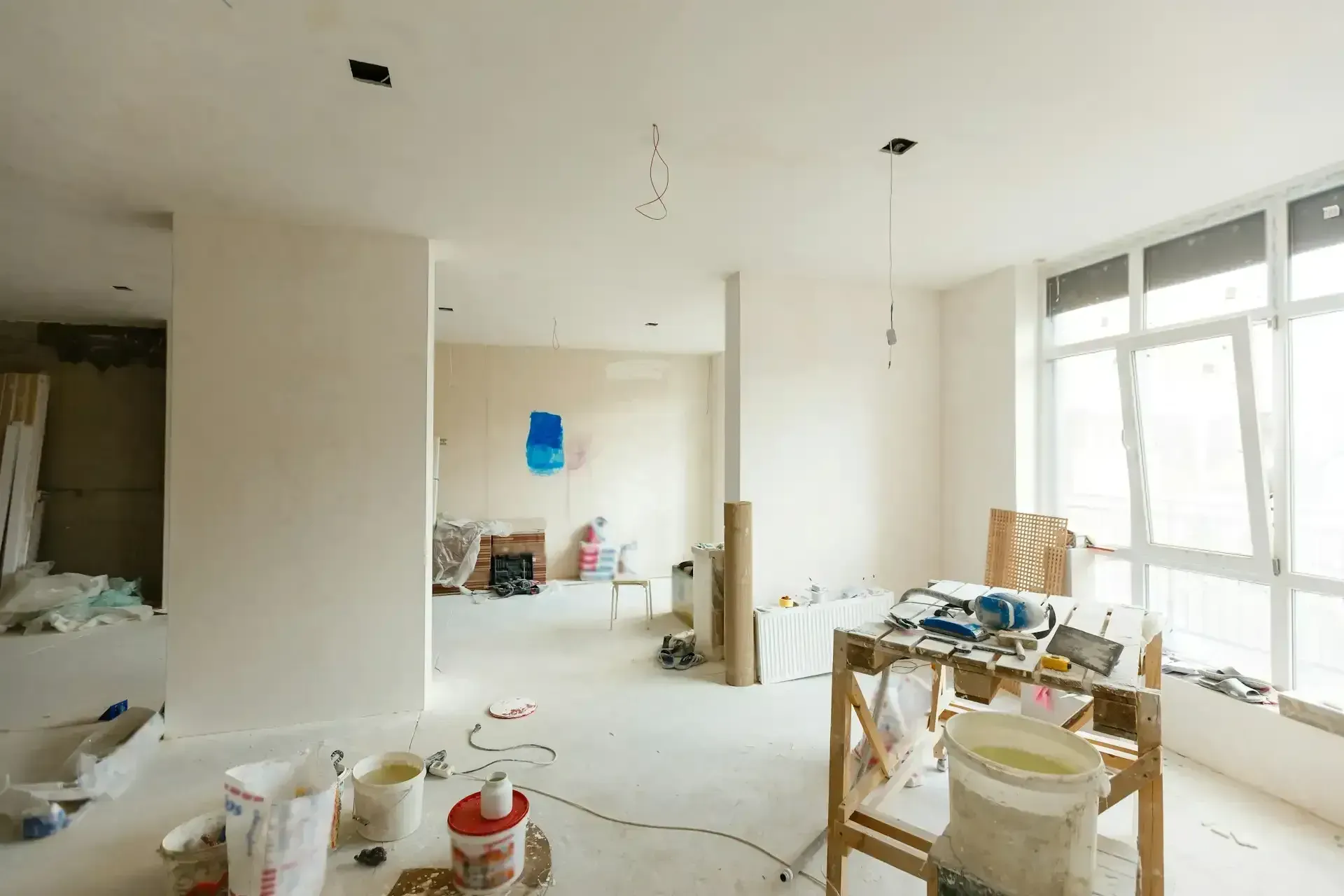 Bright unfinished apartment interior with paint supplies, tools, and large windows letting in daylight
