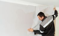 Man hanging wallpaper, smoothing a light gray wall with a tool in a bright room