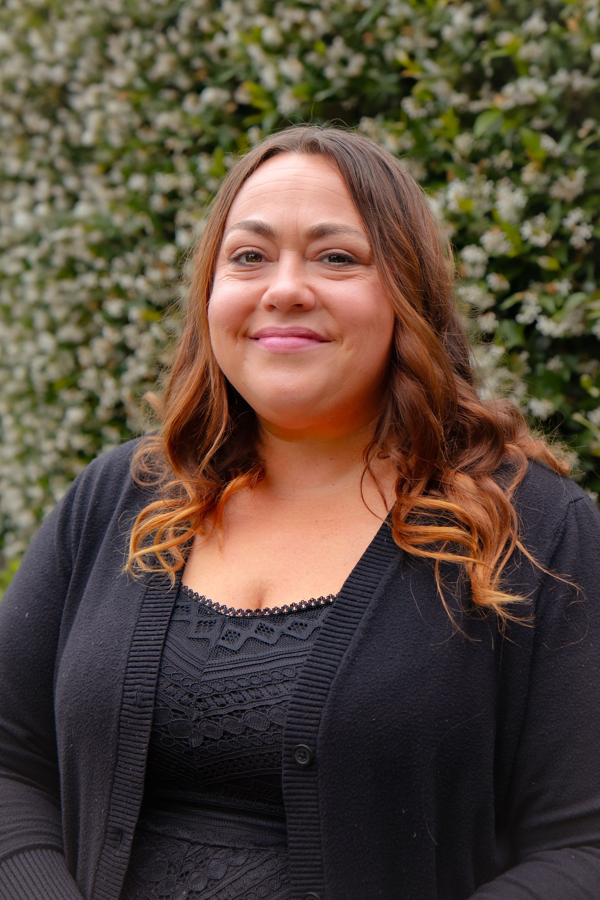 Woman with brown hair smiles, wearing a black cardigan and top, in front of green foliage.