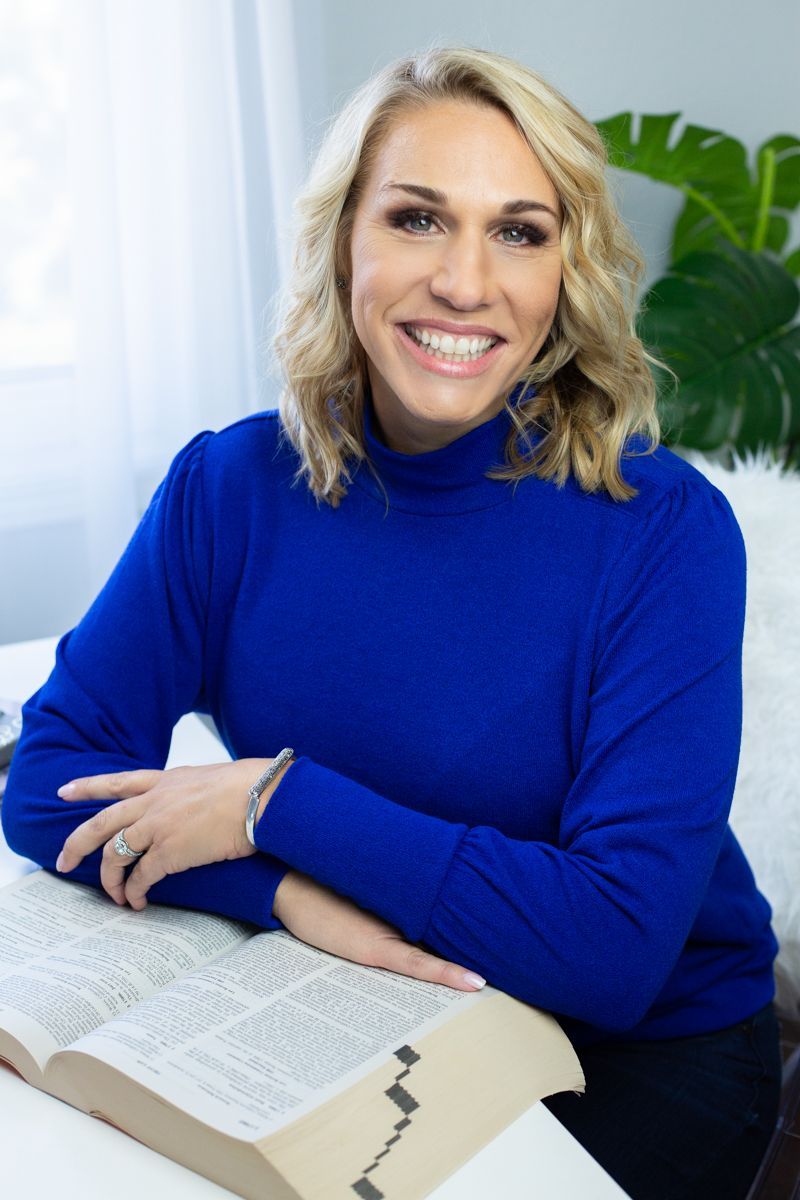 Woman with blonde hair smiles, wearing a blue sweater, leaning on an open book.
