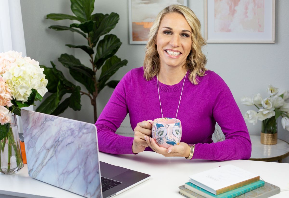 Woman in purple sweater smiles, holds mug at desk with laptop, flowers, and notebooks.