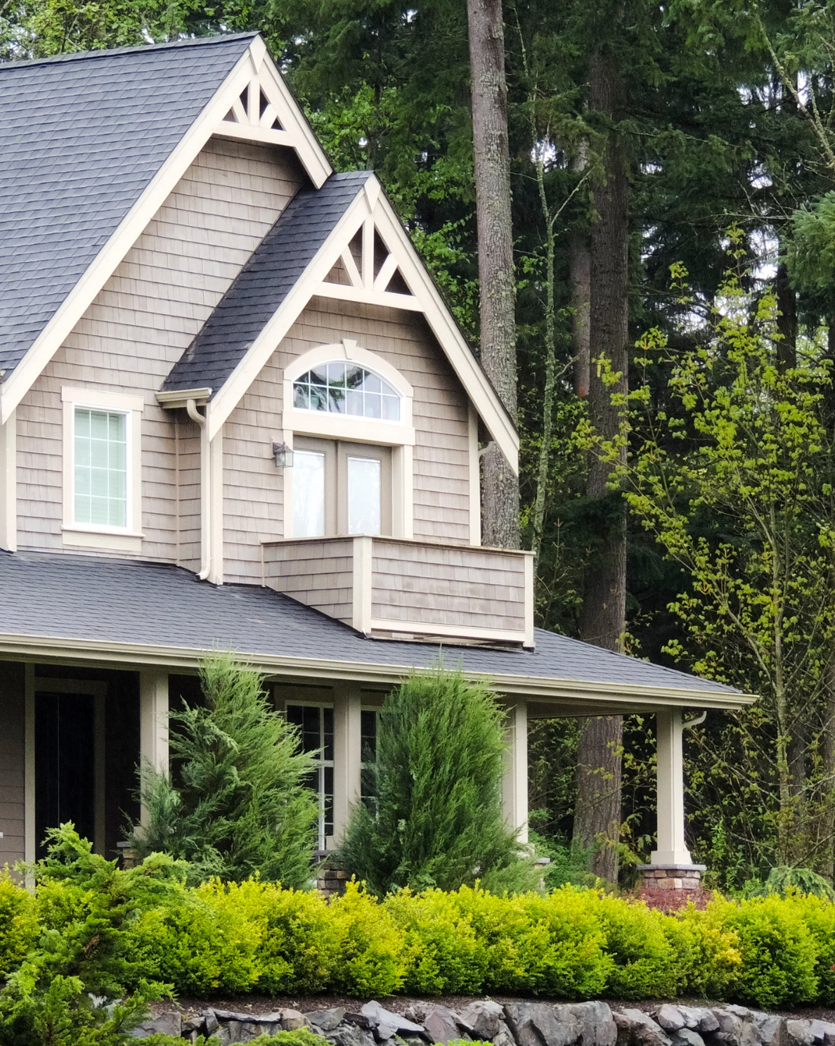 Beige two-story house with dark roof, small balcony, and porch; surrounded by trees and bushes.