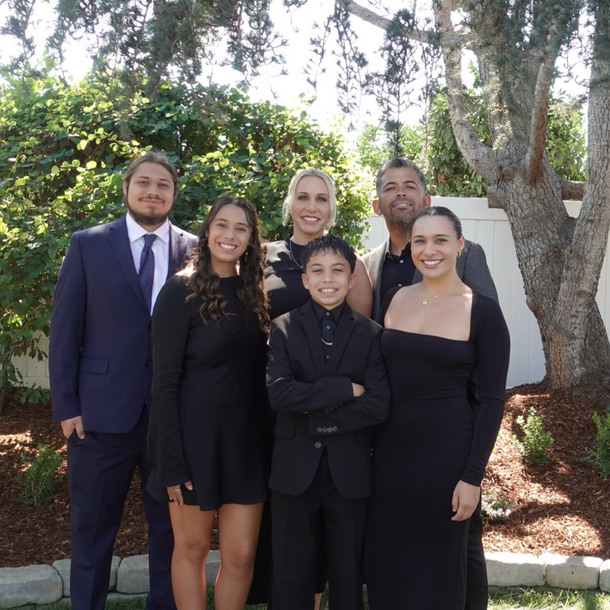Family posing outdoors: adults and children in formal attire, smiling. Lush greenery in background.