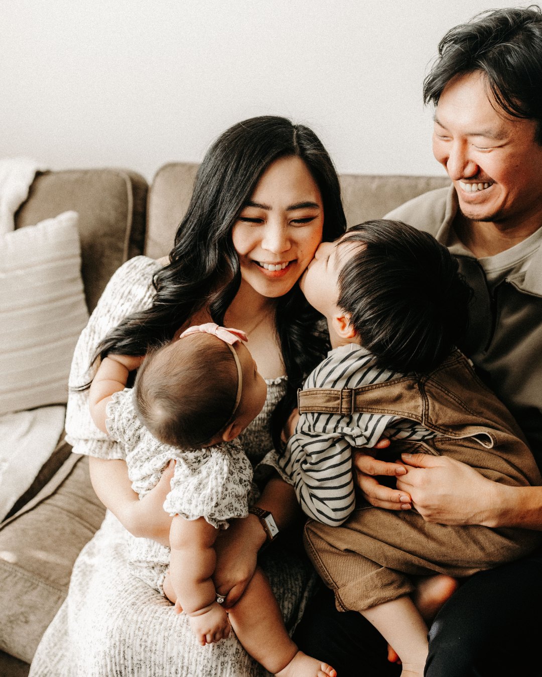 Happy family on a couch: mother holding baby, toddler kissing mother, father smiling.