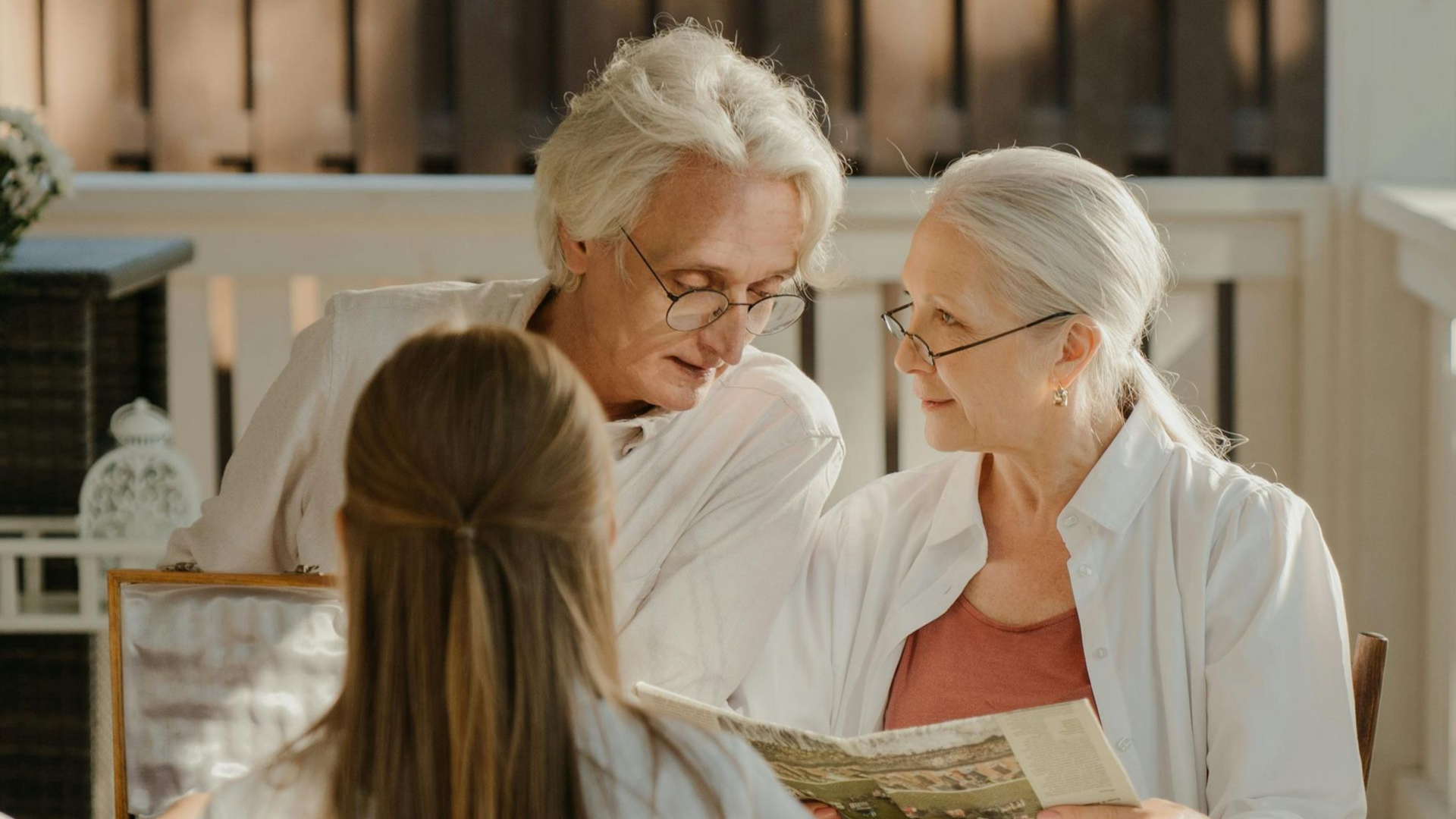 Elderly couple looking at photo album with young woman on a porch, wearing white and soft colors.
