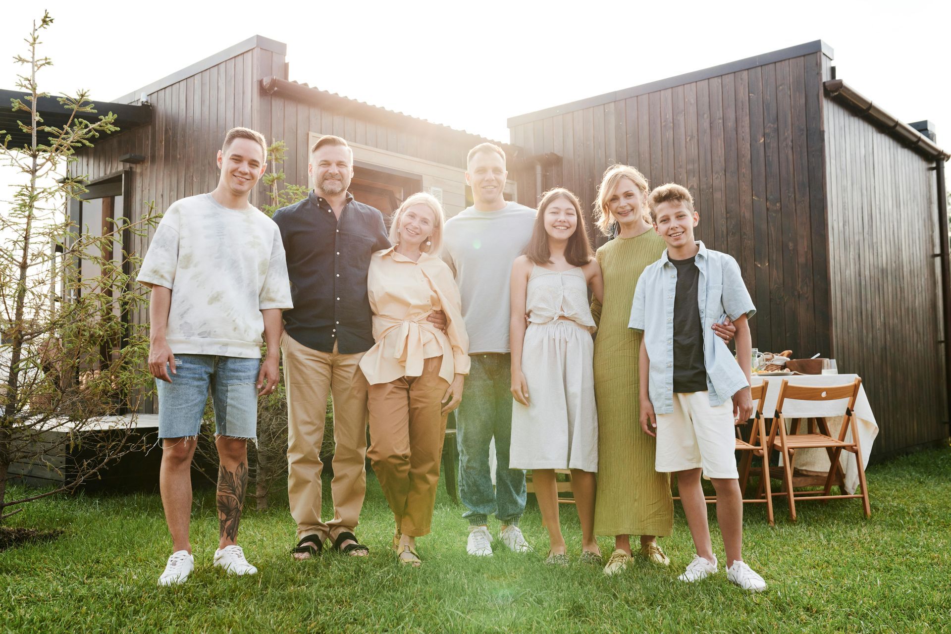 Family posing outside a wooden house on a sunny day. Smiling, green lawn, table set in the background.