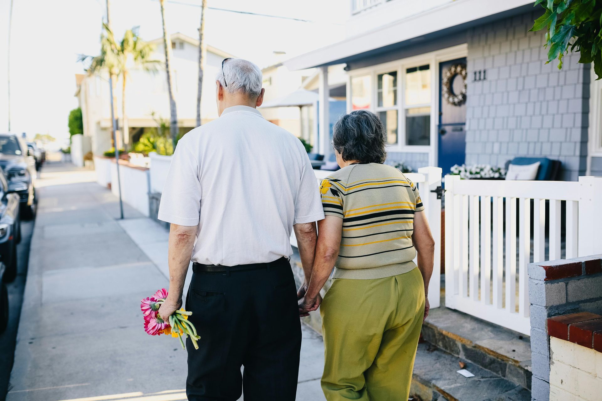 Elderly couple walks down sidewalk, holding hands. Man holds flowers. Near house.