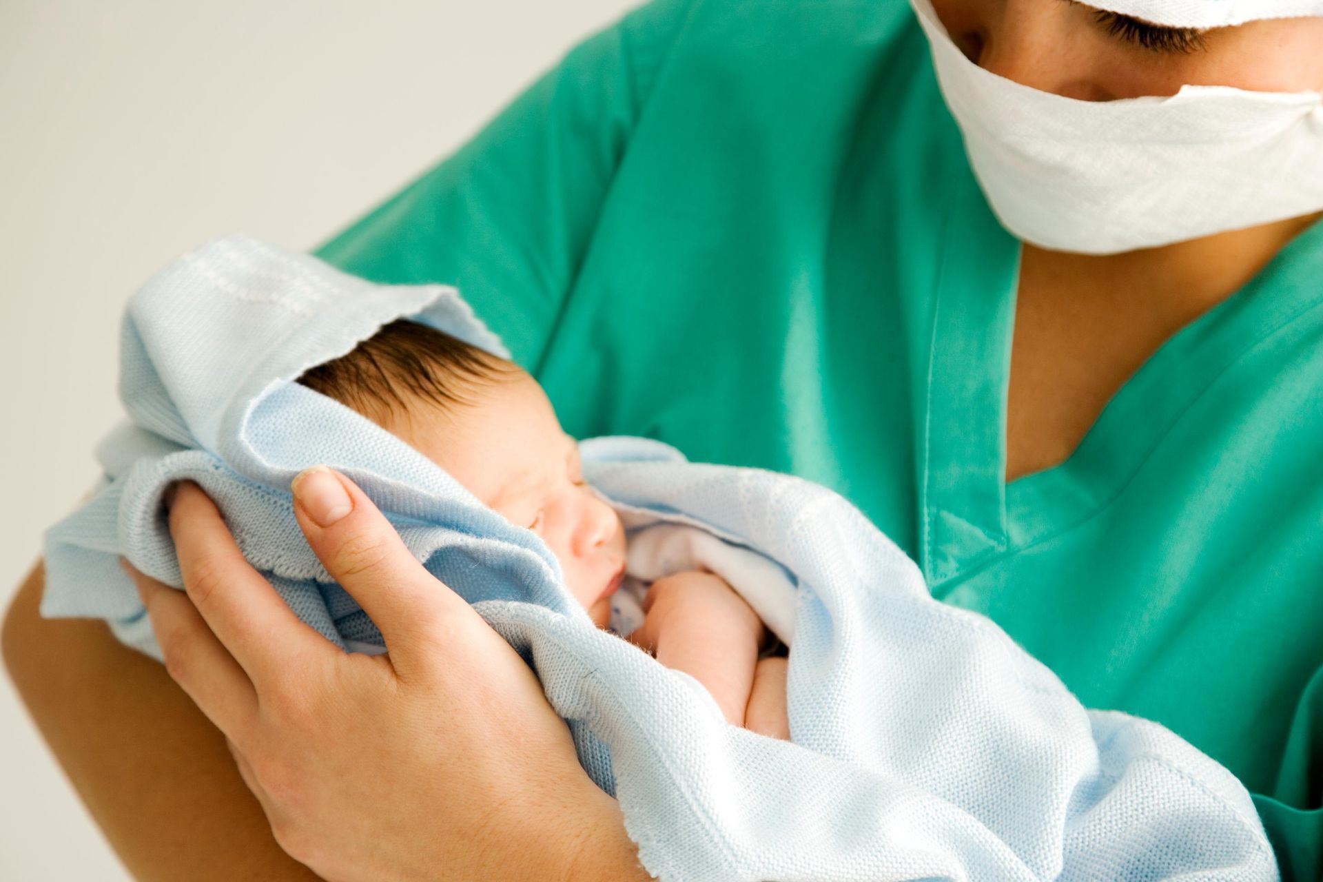 Newborn wrapped in a white blanket, held by a person in green medical scrubs and a face mask
