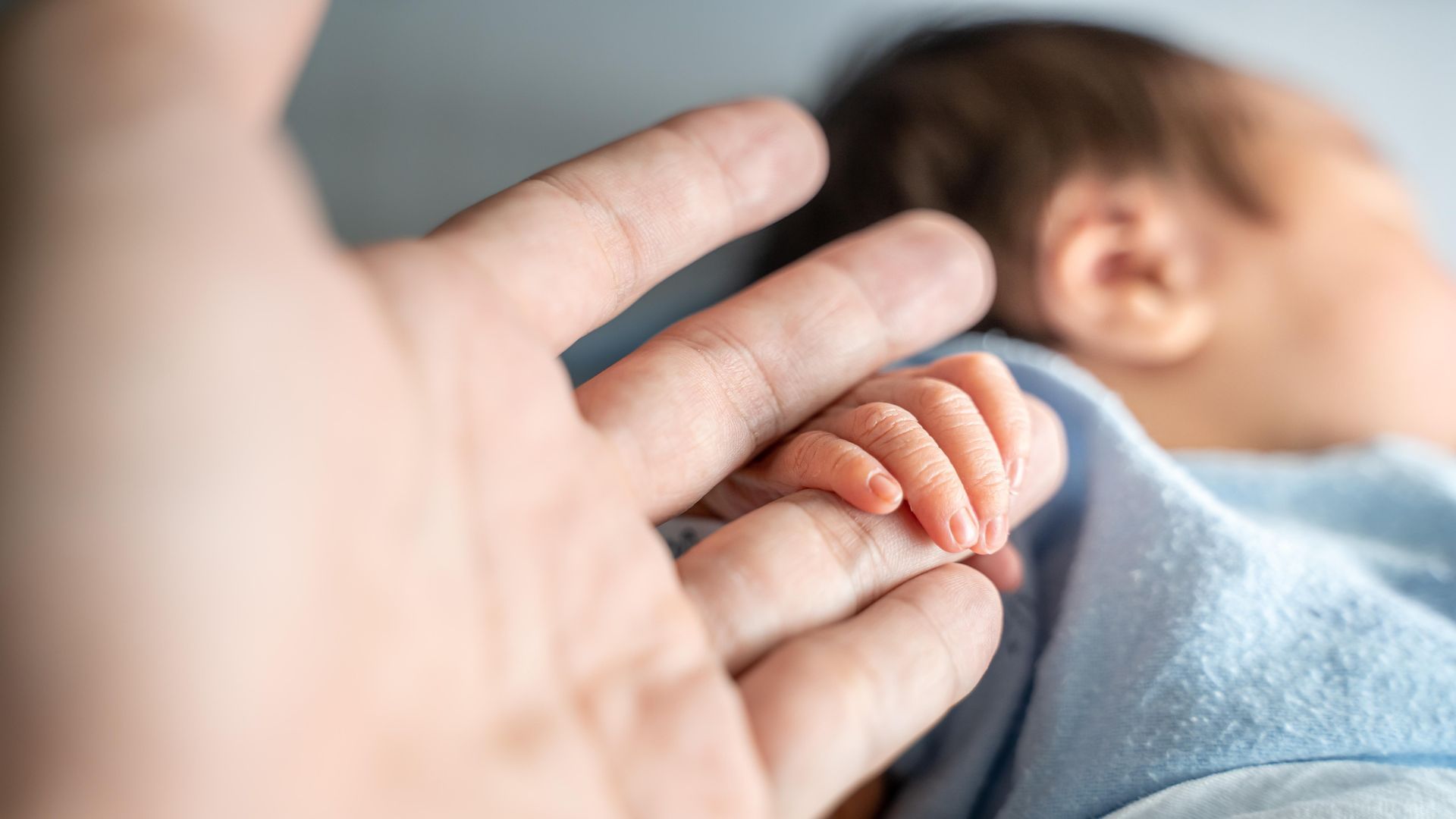 Adult hand holding a baby’s hand, with the baby lying on a blanket in the background