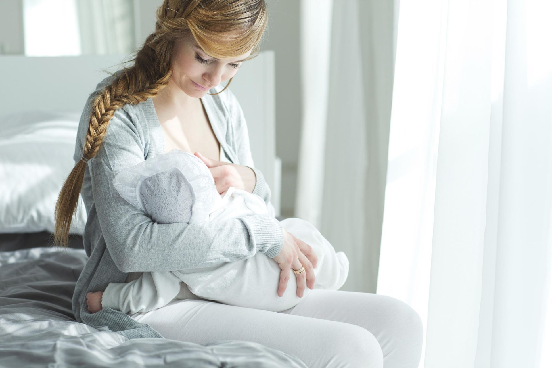 Woman breastfeeding a baby on a bed beside a bright window
