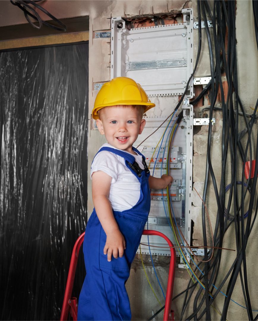 A little boy wearing a yellow hard hat and blue overalls is standing on a ladder.