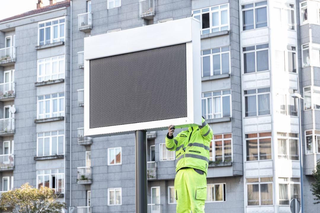 A man in a yellow jacket is holding a large billboard in front of a building.