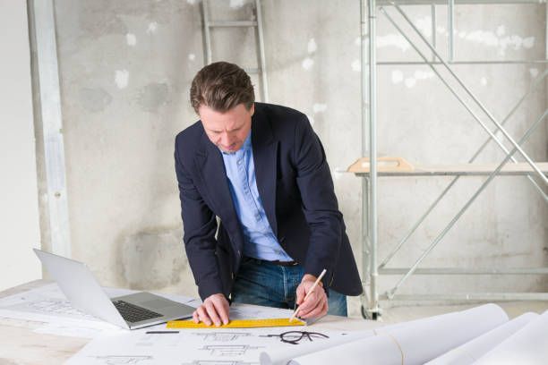 Man in blazer, werkend aan blauwdrukken met laptop en gereedschap, op de achtergrond een bouwplaats.