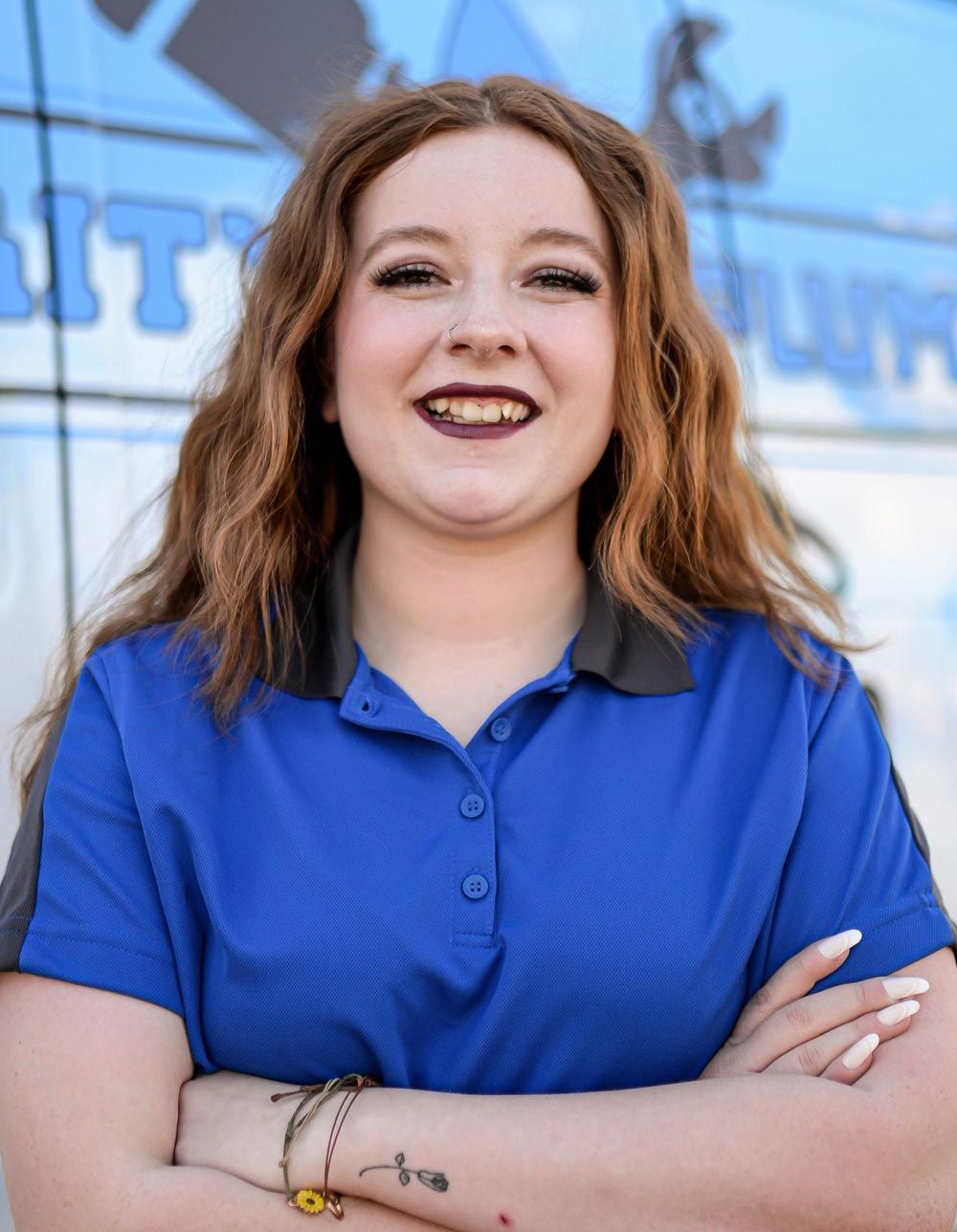 Young woman with reddish-brown hair, arms crossed, wearing blue polo shirt smiles at the camera outdoors.