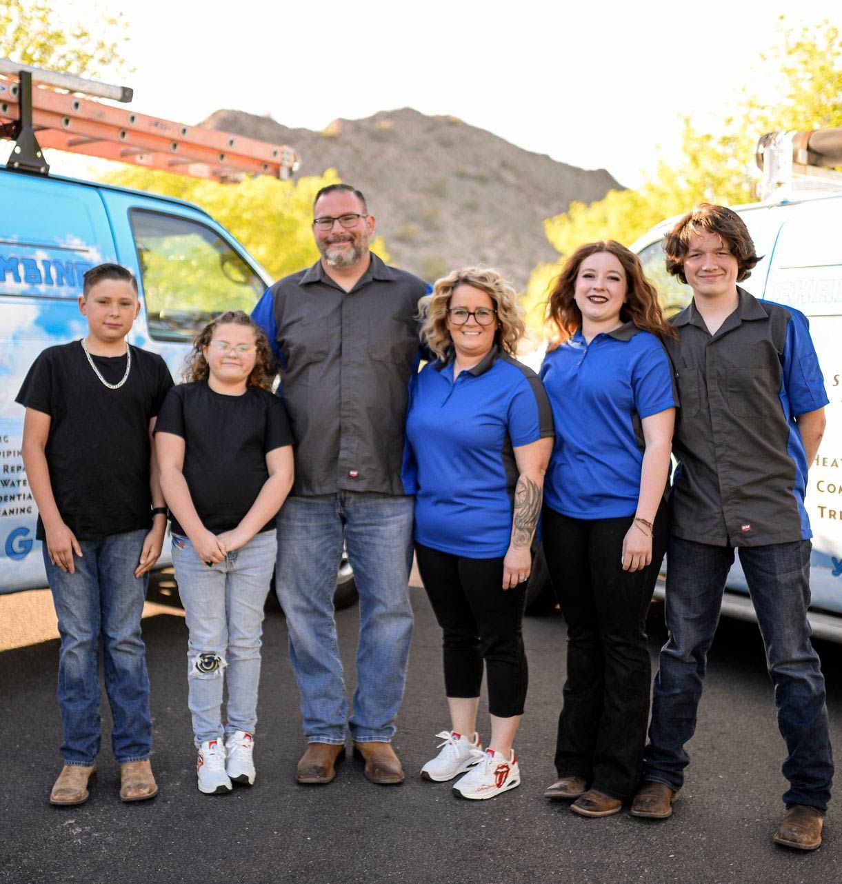 Family in front of work vans. Man, woman and four children smiling outside, mountains in background.