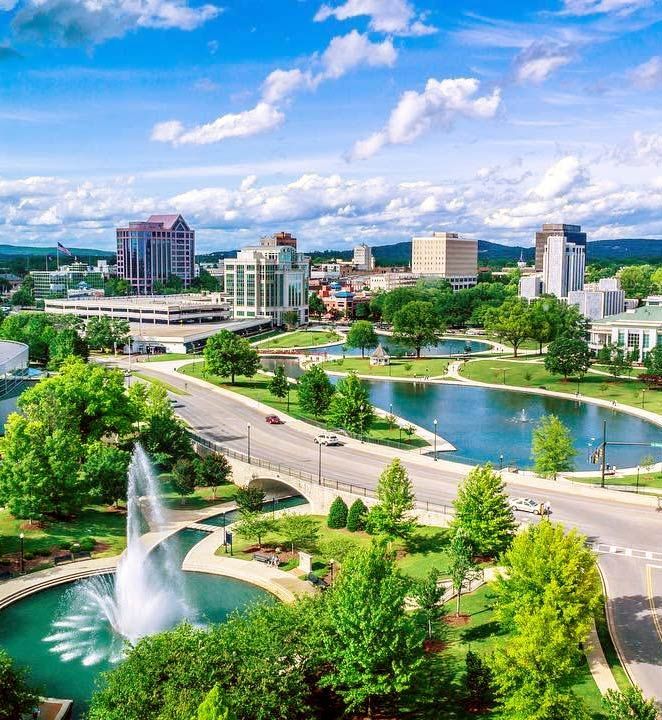 Cityscape with buildings, water features, green trees, and a bright blue sky.