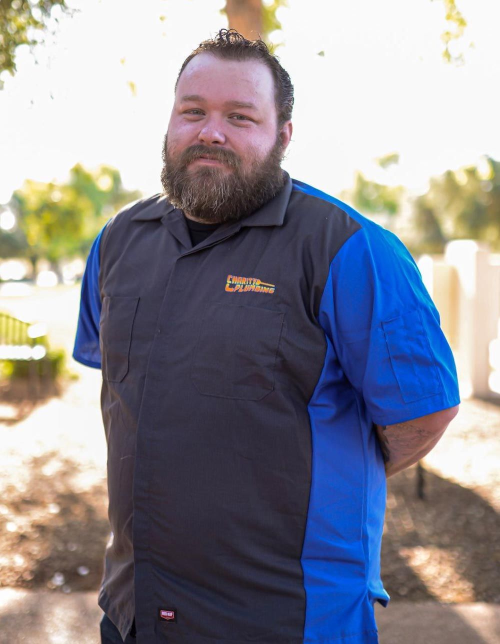 Man with a beard wearing a blue and grey work shirt, standing outside in sunlight.