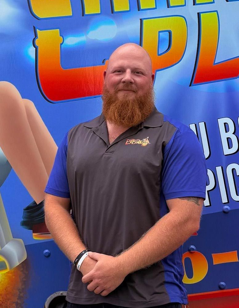 Bald man with a red beard wearing a blue and gray polo shirt, in front of a sign with colorful text.