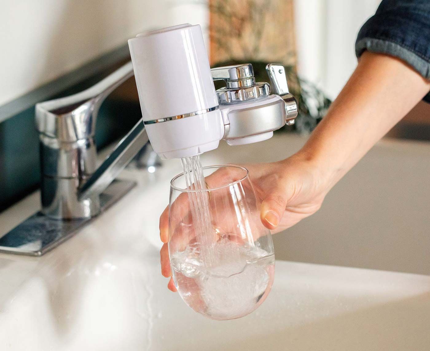 Person filling a glass of water from a white water filter attached to a kitchen faucet.