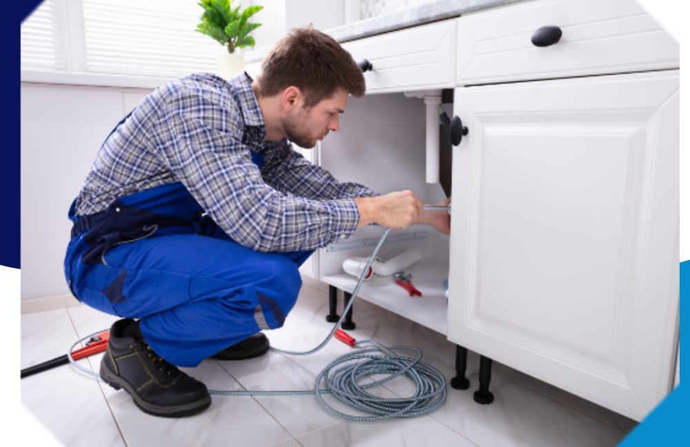Man in blue overalls working under a white kitchen cabinet, using a cable.