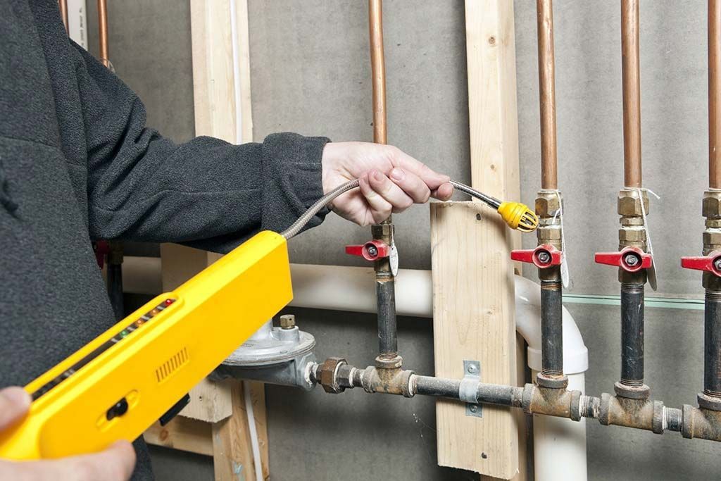 Person uses a yellow gas leak detector to inspect copper pipes with red valves.