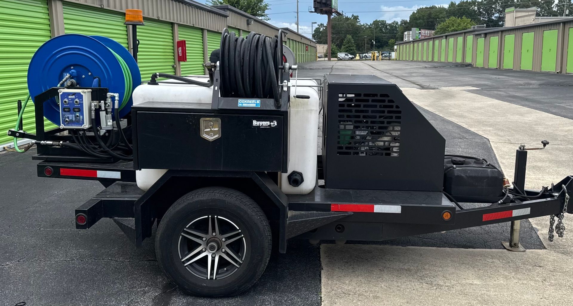 Black trailer with blue hose reel, water tank, and generator, parked in front of storage units.