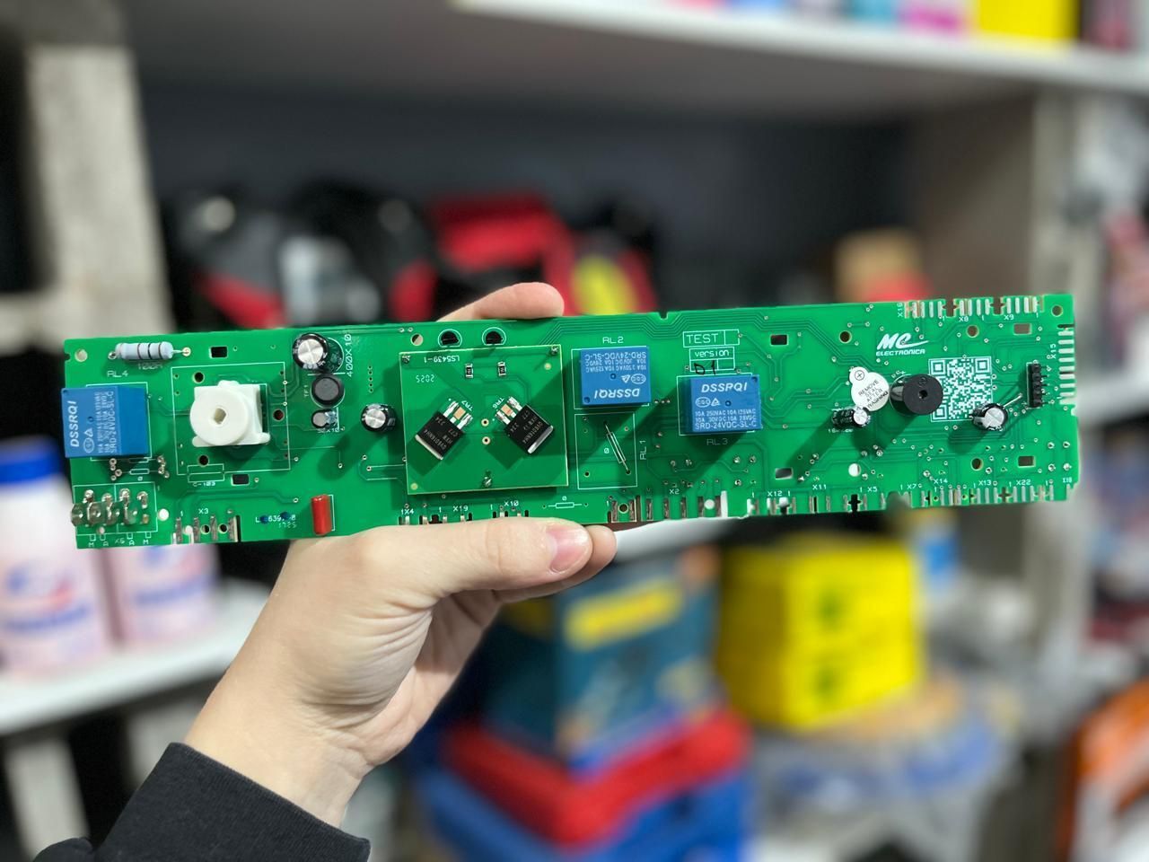 Una mano sostiene una larga placa de circuito verde en un taller, con estantes borrosos al fondo.
