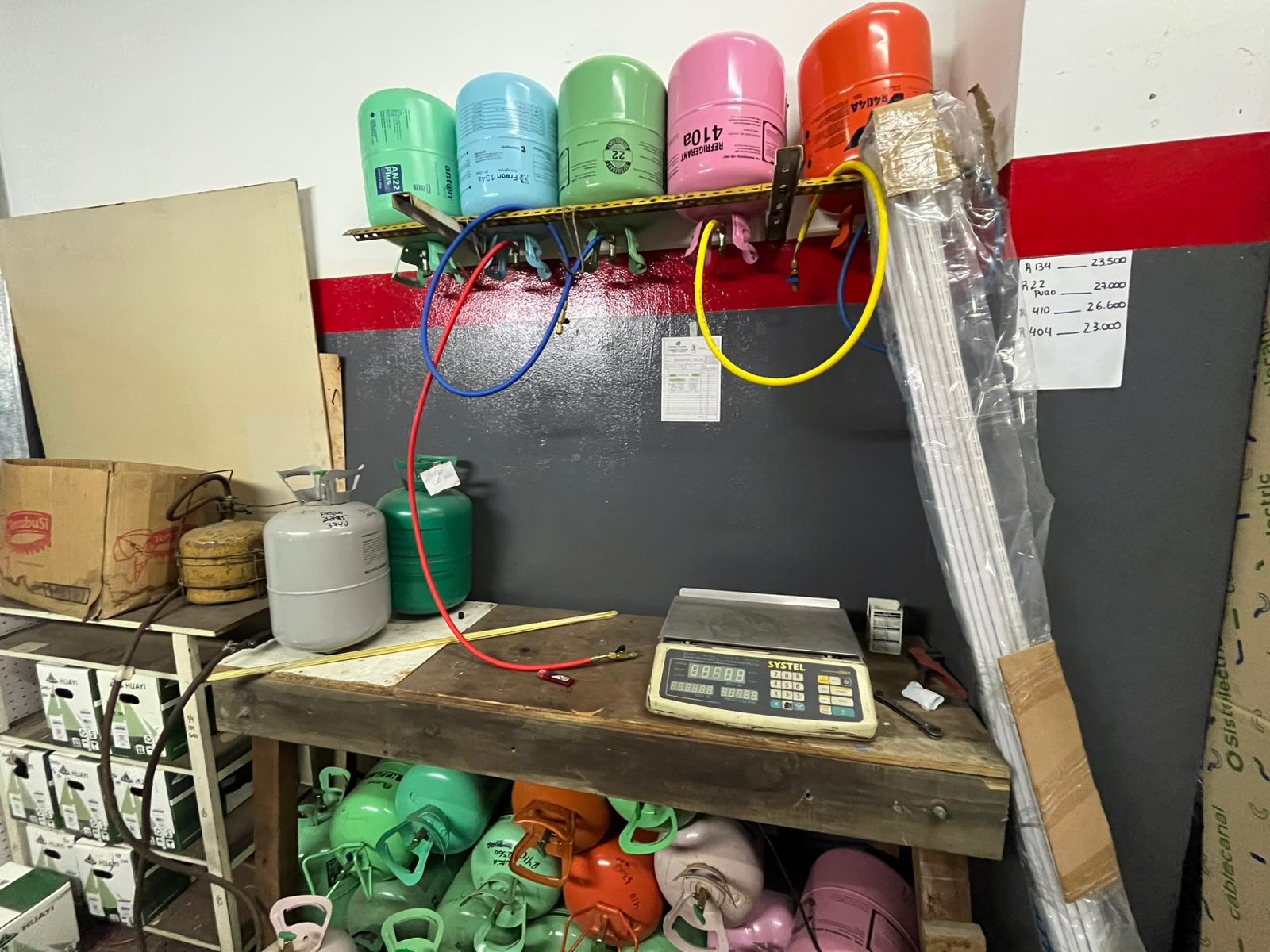 Tanques y tuberías industriales codificados por colores, montados en la pared de un taller, con herramientas y piezas en estantes debajo.