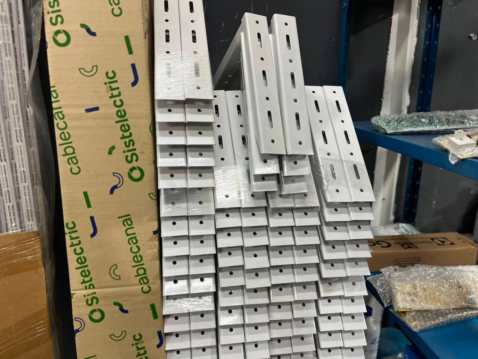 Rieles metálicos blancos apilados y soportes perforados almacenados en estanterías de almacén junto a una caja de cartón.