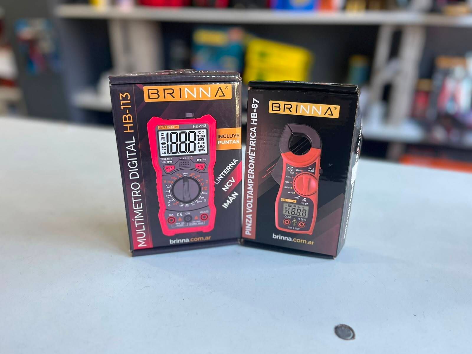 Dos multímetros digitales BRINNA en cajas de color negro y naranja sobre una mesa, con una moneda delante.