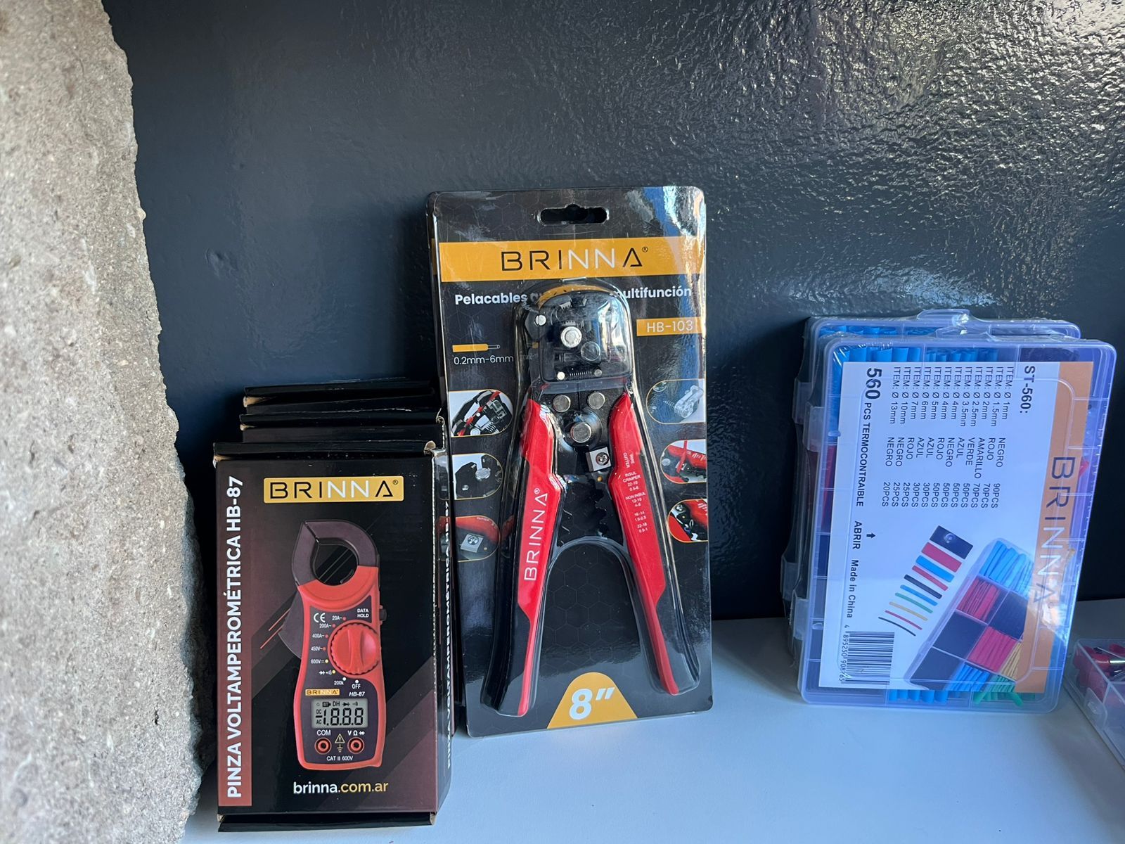 Tres herramientas en cajas sobre un estante: un medidor negro, unos alicates rojos y un estuche de plástico azul.