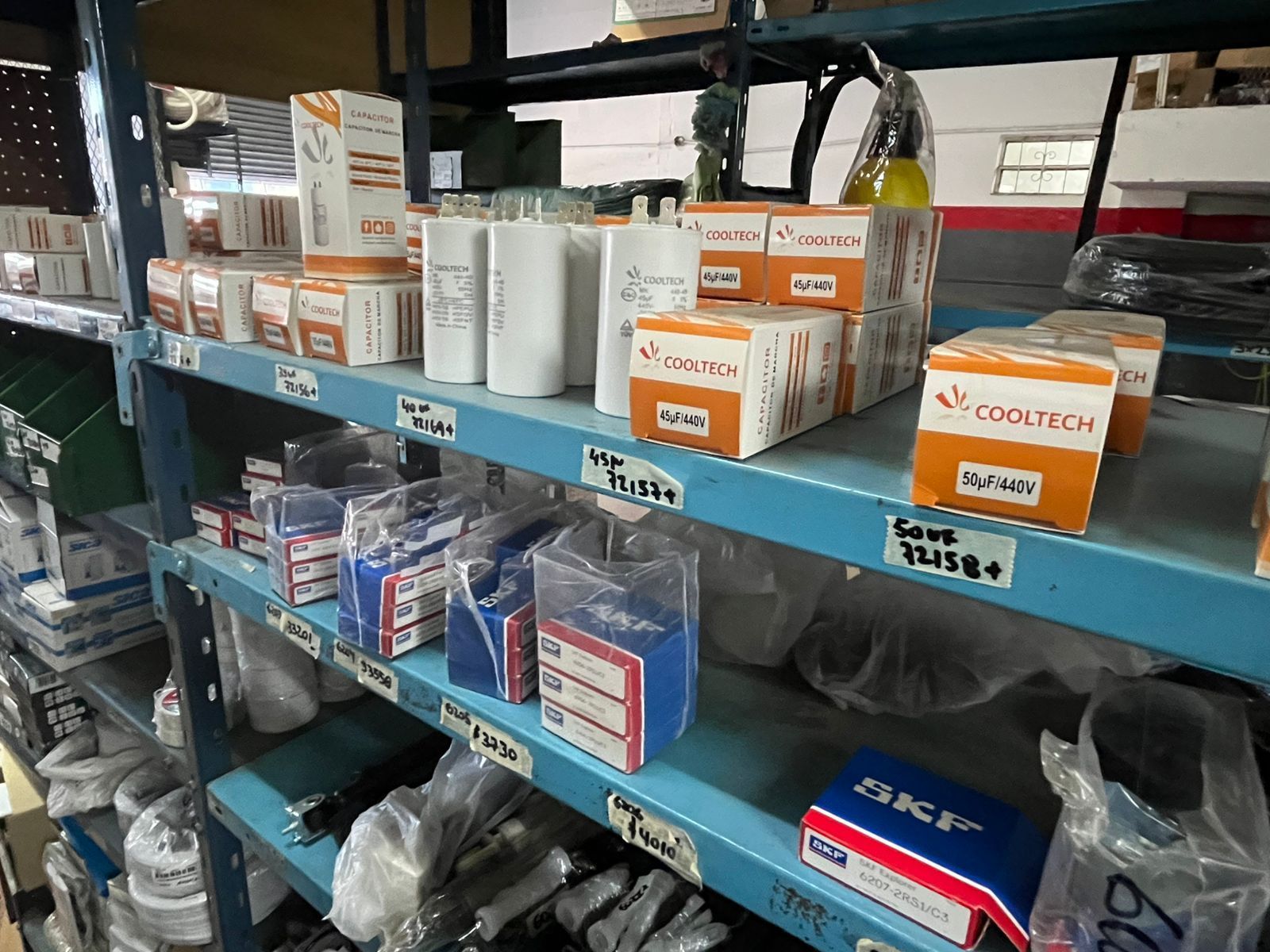 Estanterías de almacén repletas de piezas variadas, empaquetadas en cajas y bolsas; las etiquetas son visibles en los estantes metálicos azules.
