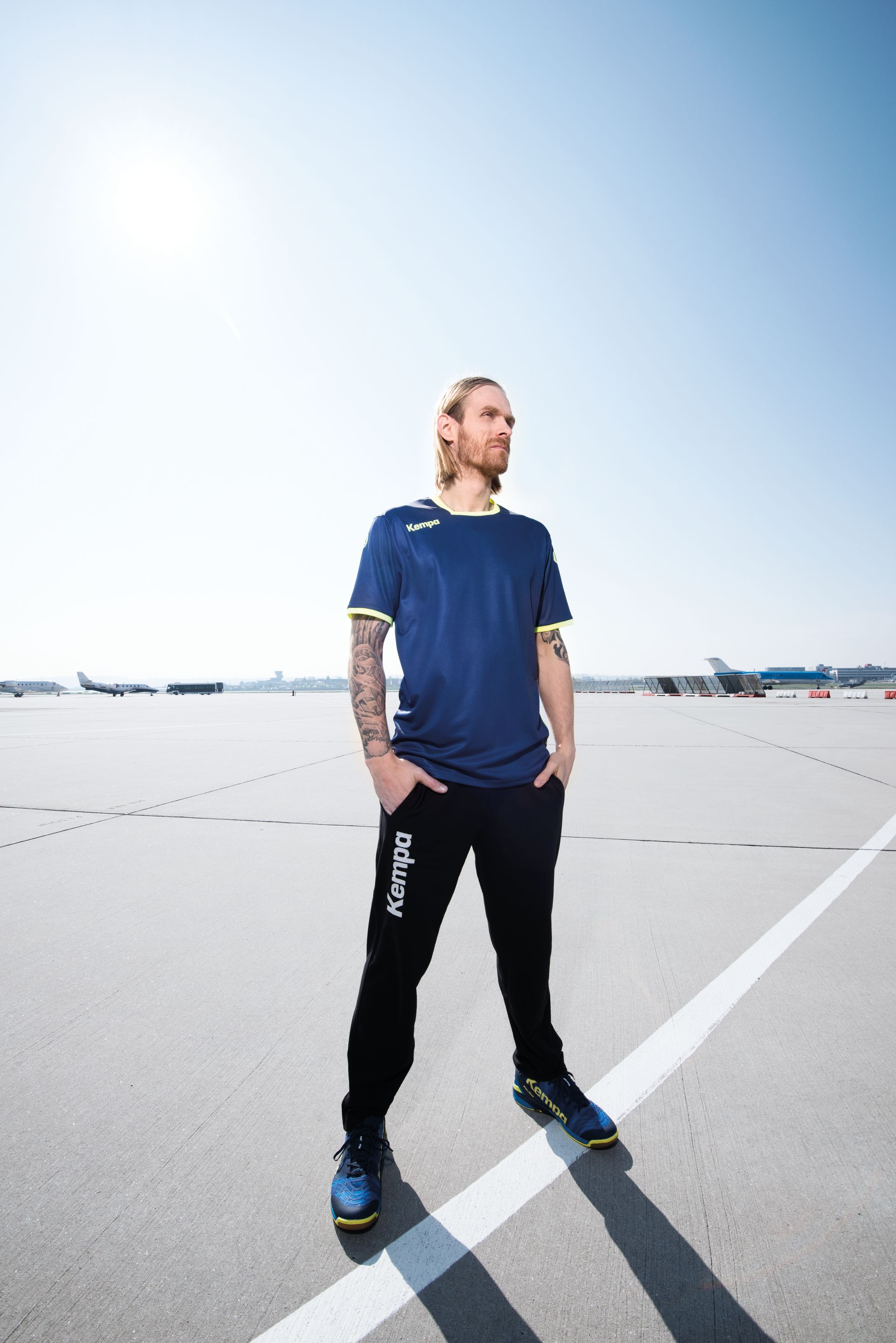 Maður með sítt hár, bláa skyrtu og svartar buxur stendur á flugbraut og horfir til hliðar.