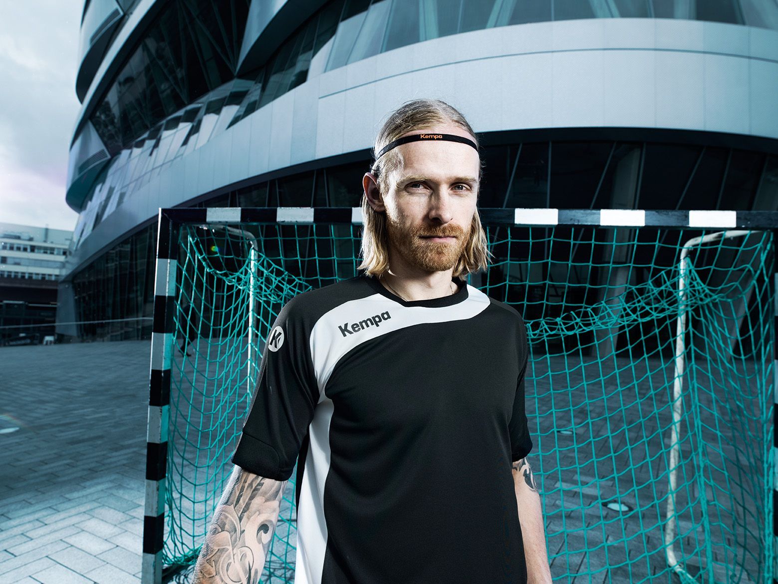 Handboltamaður með skegg fyrir framan markið, nútímaleg bygging í bakgrunni.