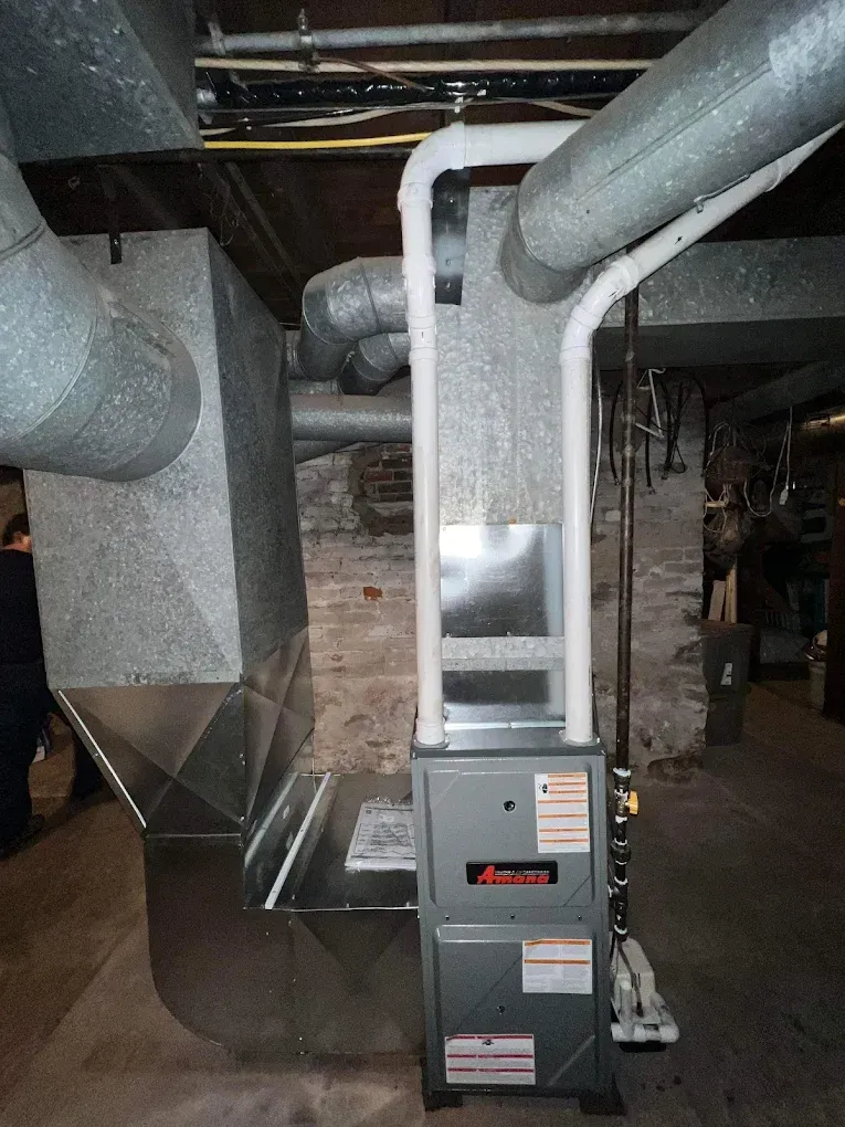 Furnace unit with ductwork in a basement setting. Grey metal and brick walls.