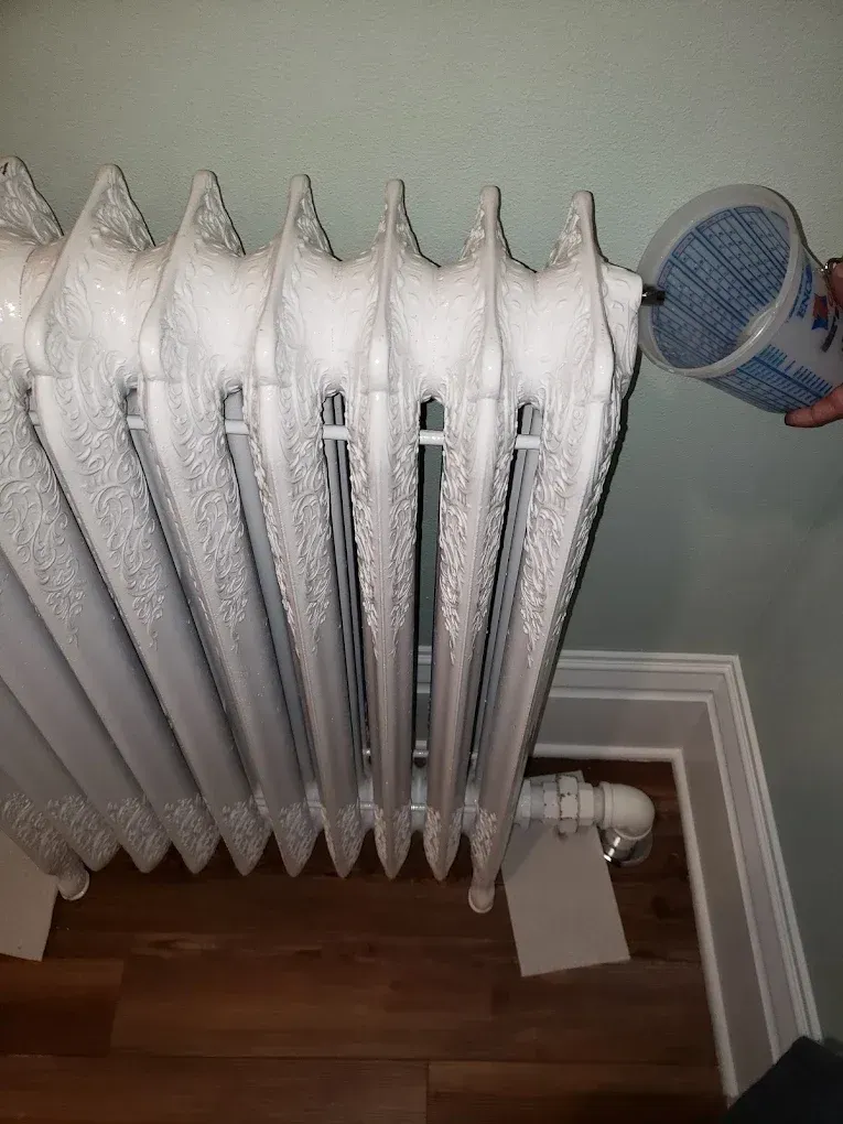 Person pours blue-tinged liquid onto a white radiator with ornate detailing, in a room with hardwood floors.