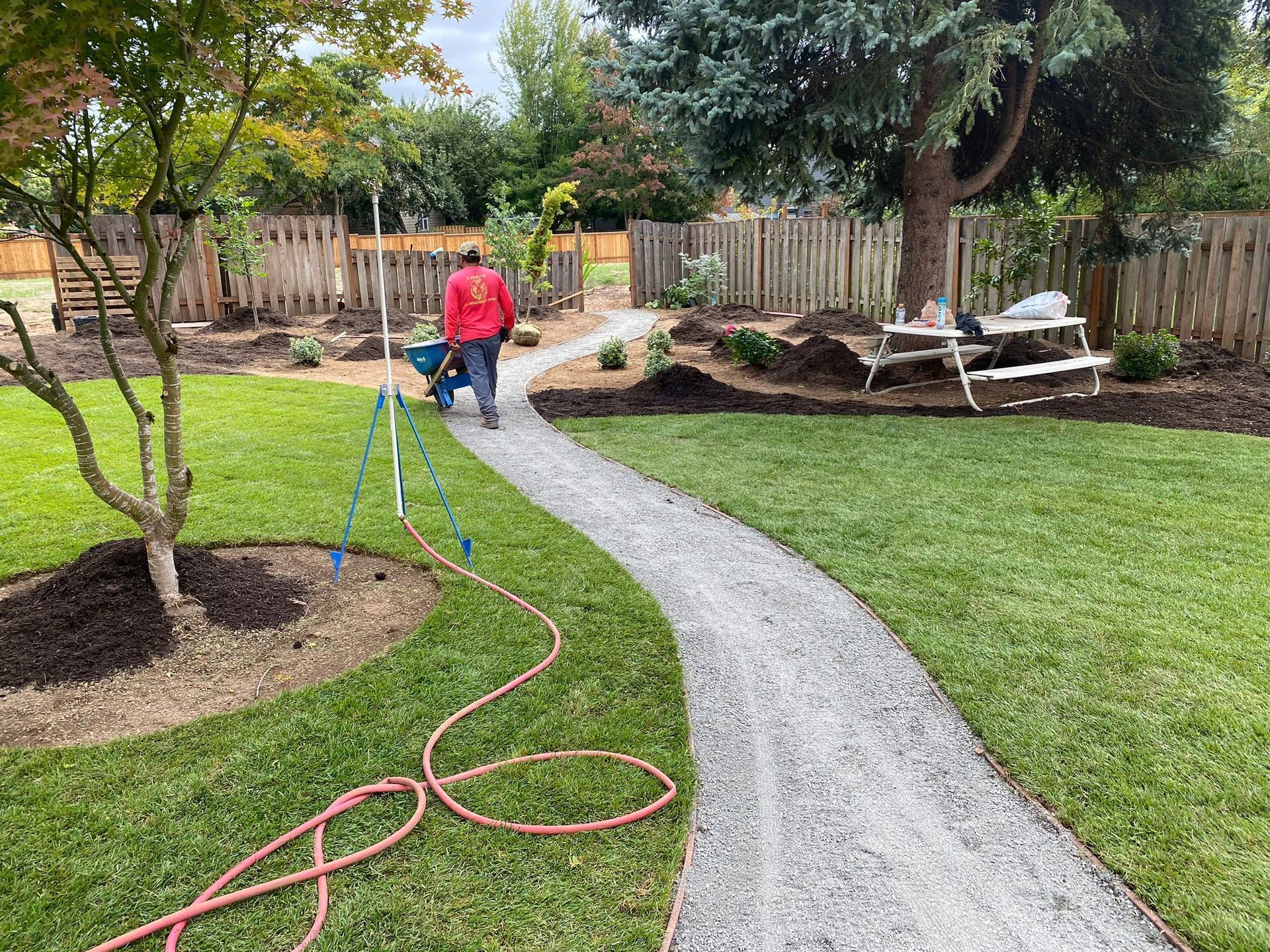 Person pushing a wheelbarrow on a gravel path in a garden. Mulch beds, lawn, and wooden fence visible.