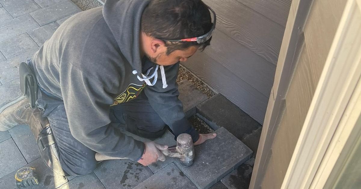 Person kneeling, hammering a stone tile near a building, wearing a hoodie and headlamp.