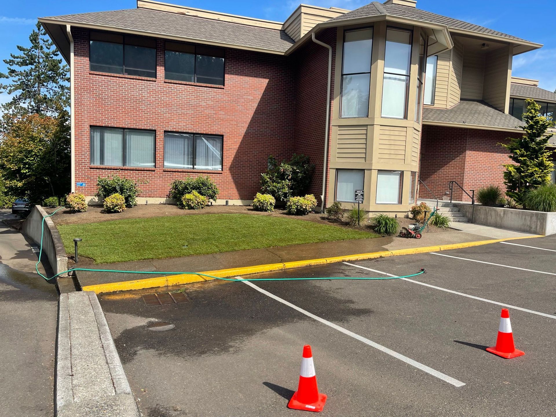 Apartment building with lawn and asphalt parking lot, water on the ground, two traffic cones.