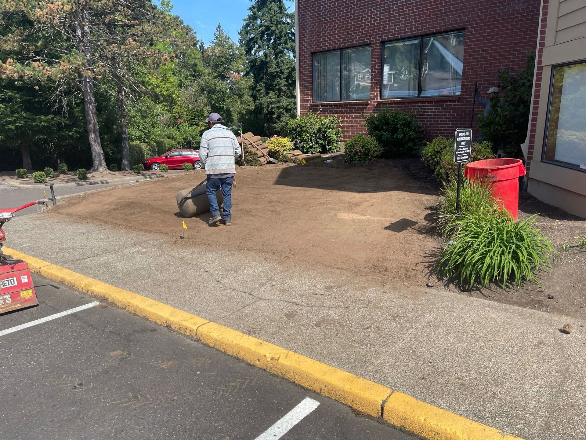 Man tilling soil in a small garden bed next to a building and a parking area with a yellow curb.