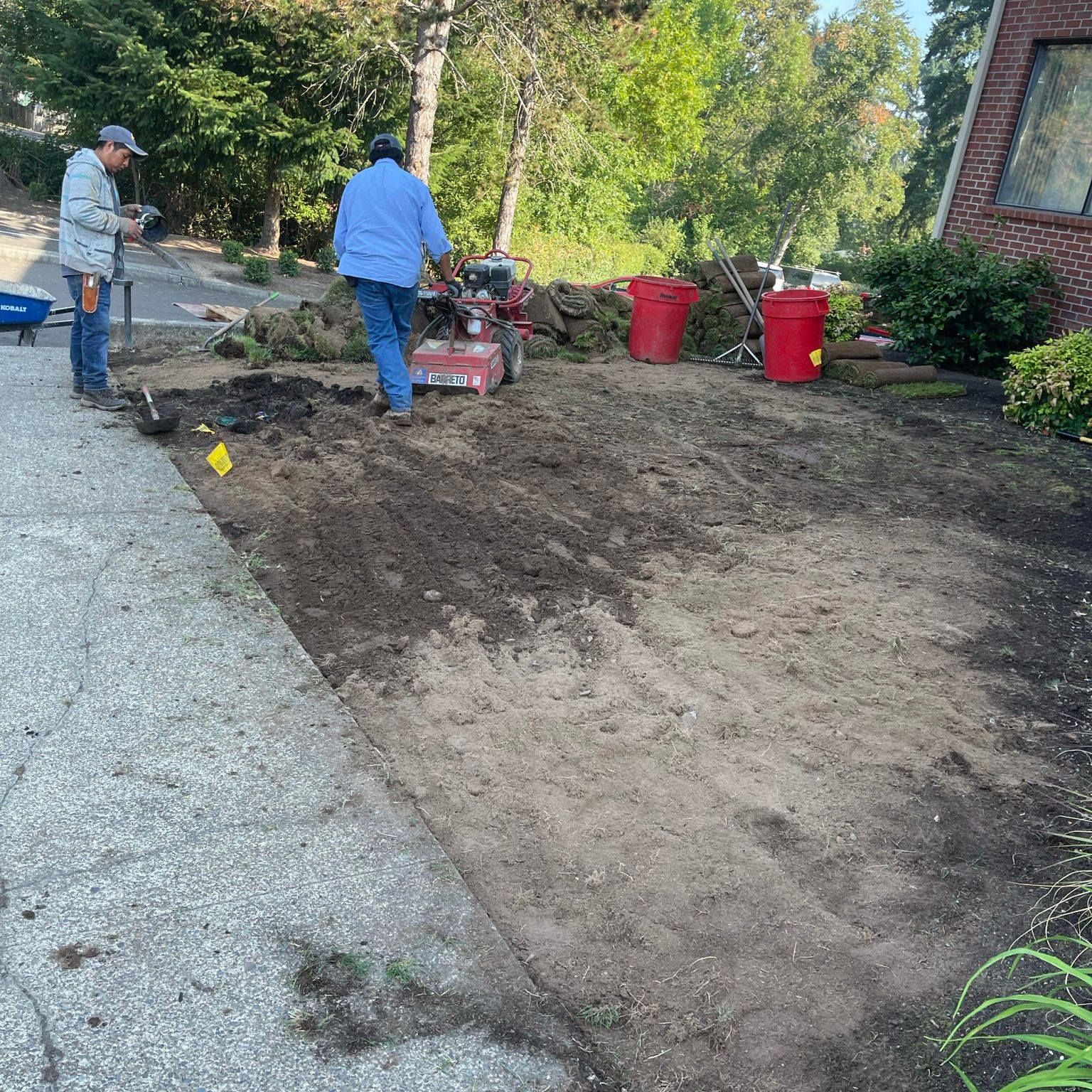 Two people tilling a yard next to a driveway; red buckets and a house in the background.