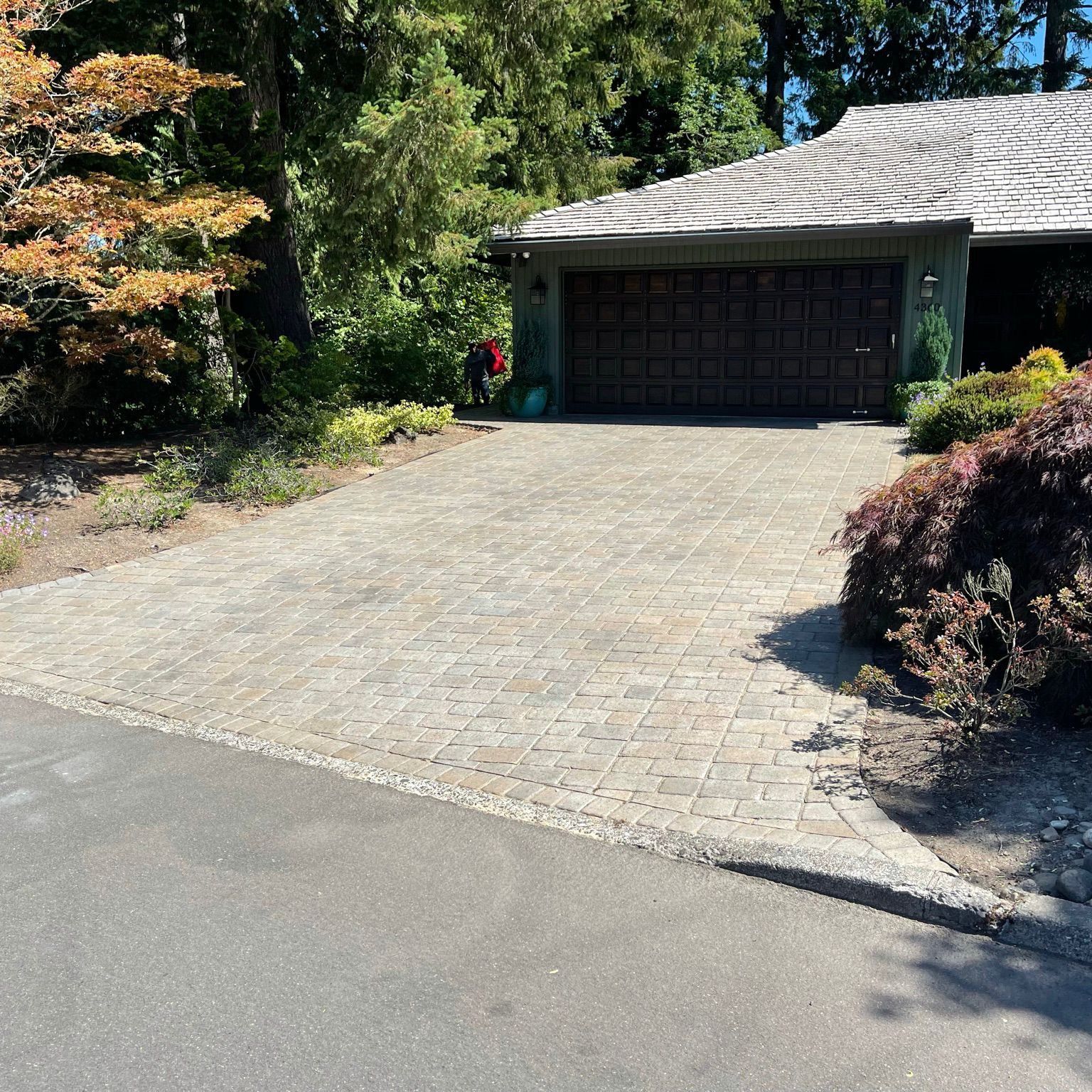 Brick driveway leading to a house with a brown garage door, trees, and shrubs on a sunny day.