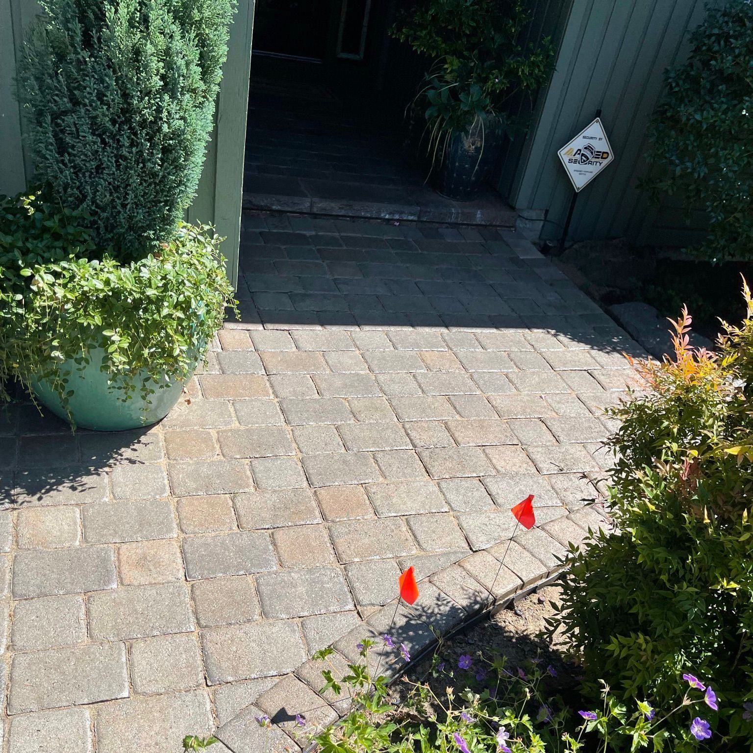 Brick walkway leading to a doorway, flanked by potted plants. Red flags mark a garden bed.