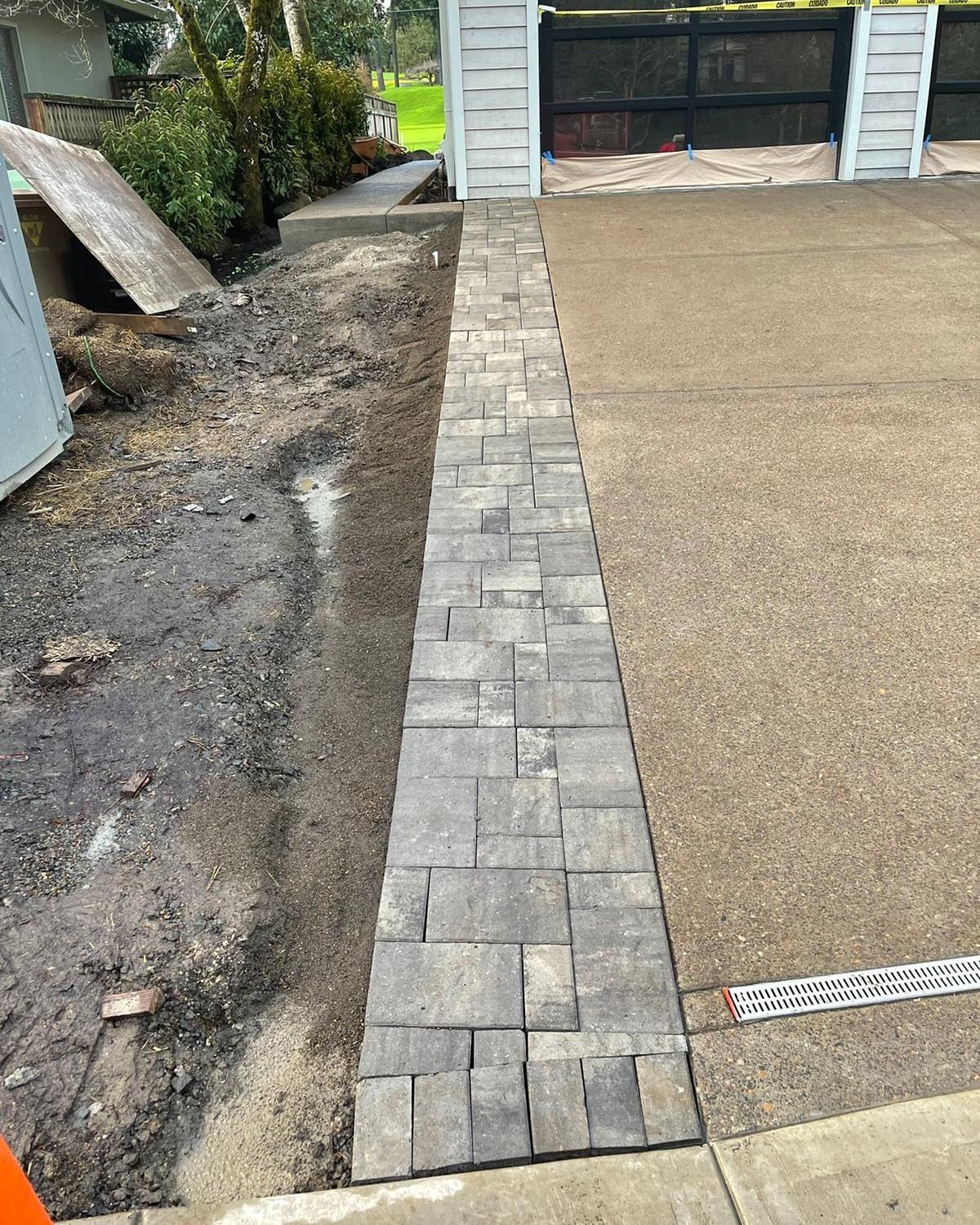 Brick pathway next to a driveway. Pathway is dark grey and leads toward a white gate.