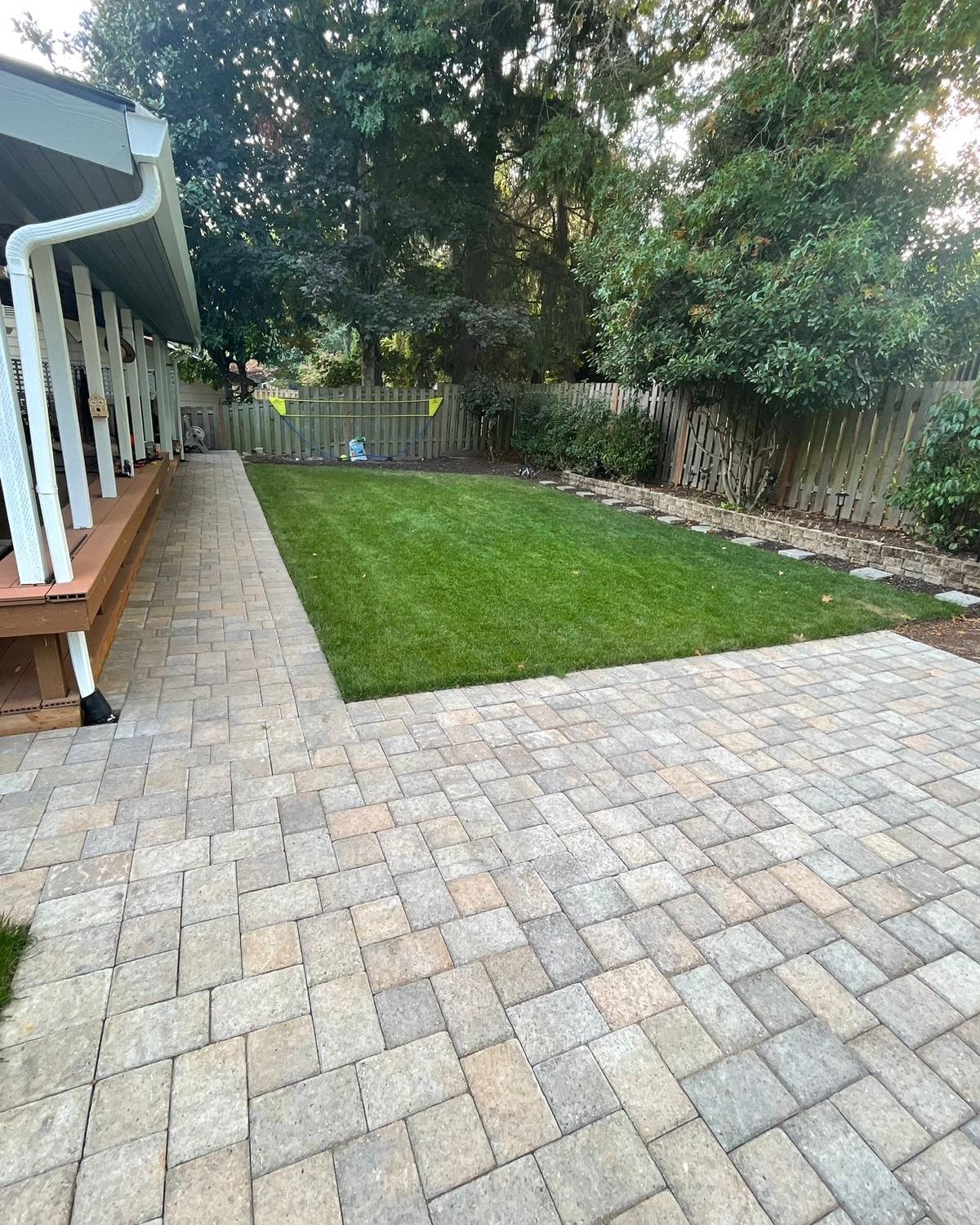 Brick patio and walkway leading to a grassy yard bordered by a wooden fence and trees.