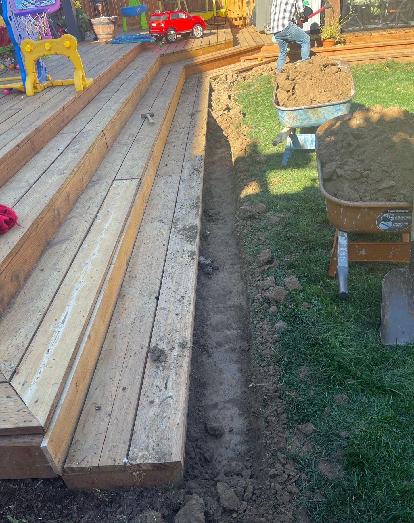 Trench dug alongside wooden deck stairs in a grassy yard, with a person filling a wheelbarrow with dirt.