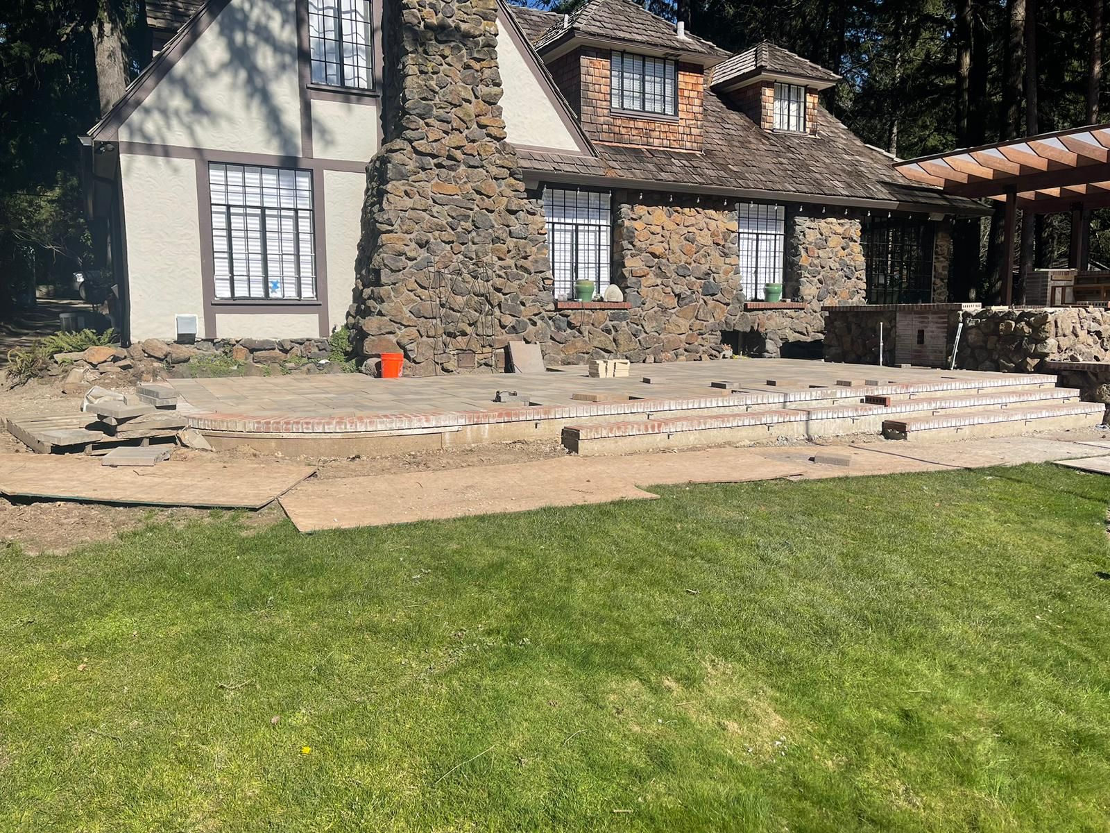 Backyard patio under construction in front of a stone house with green grass.
