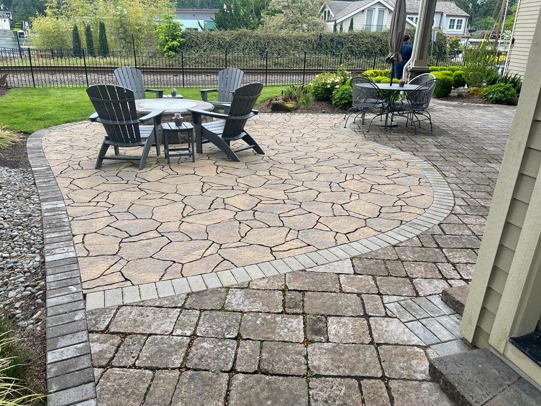 Stone patio with outdoor furniture, edged by brick. Green grass, trees, and buildings are in the background.